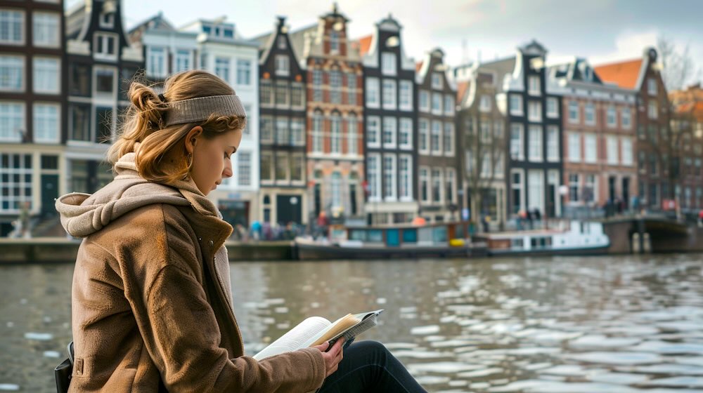 een vrouw met een donkerbruine jas leest een boek van a4 formaat naast de grachten in Amsterdam