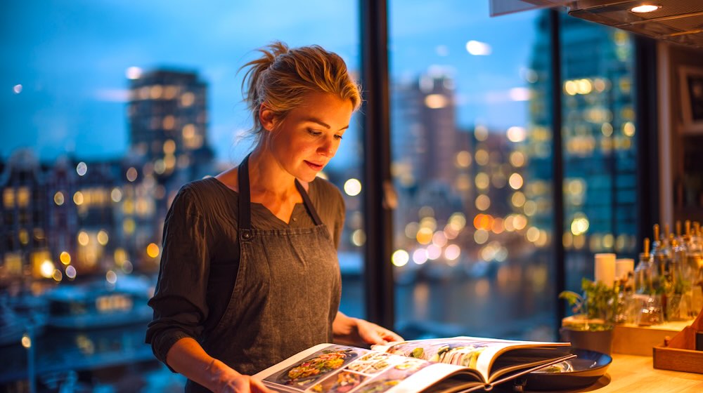 blonde vrouw in zwarte kleding gebruikt kookboek in moderne keuken in Amsterdam.