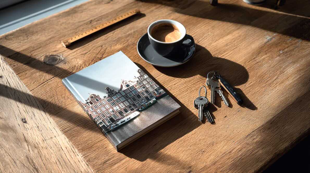 een bruine houten tafel met daarop een boek in a5 formaat en een cover van grachtenpanden in Amsterdam, naast een kop koffie en twee bosjes met sleutels