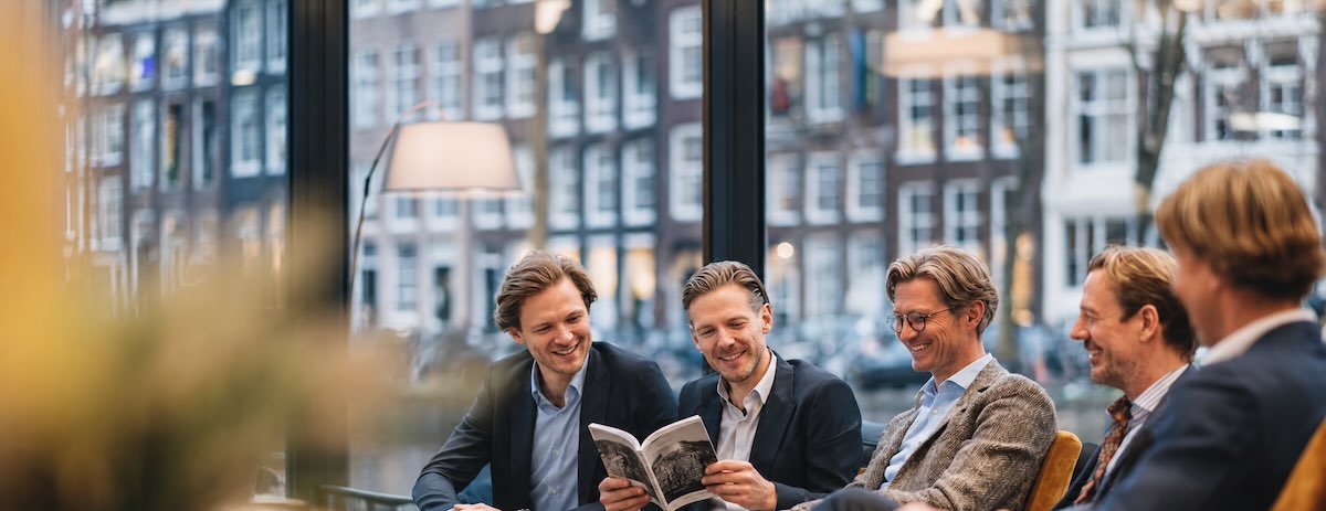 Vijf mannen lezen samen een boek in b5 formaat in kantoor met Amsterdamse grachtenpanden op de achtergrond.